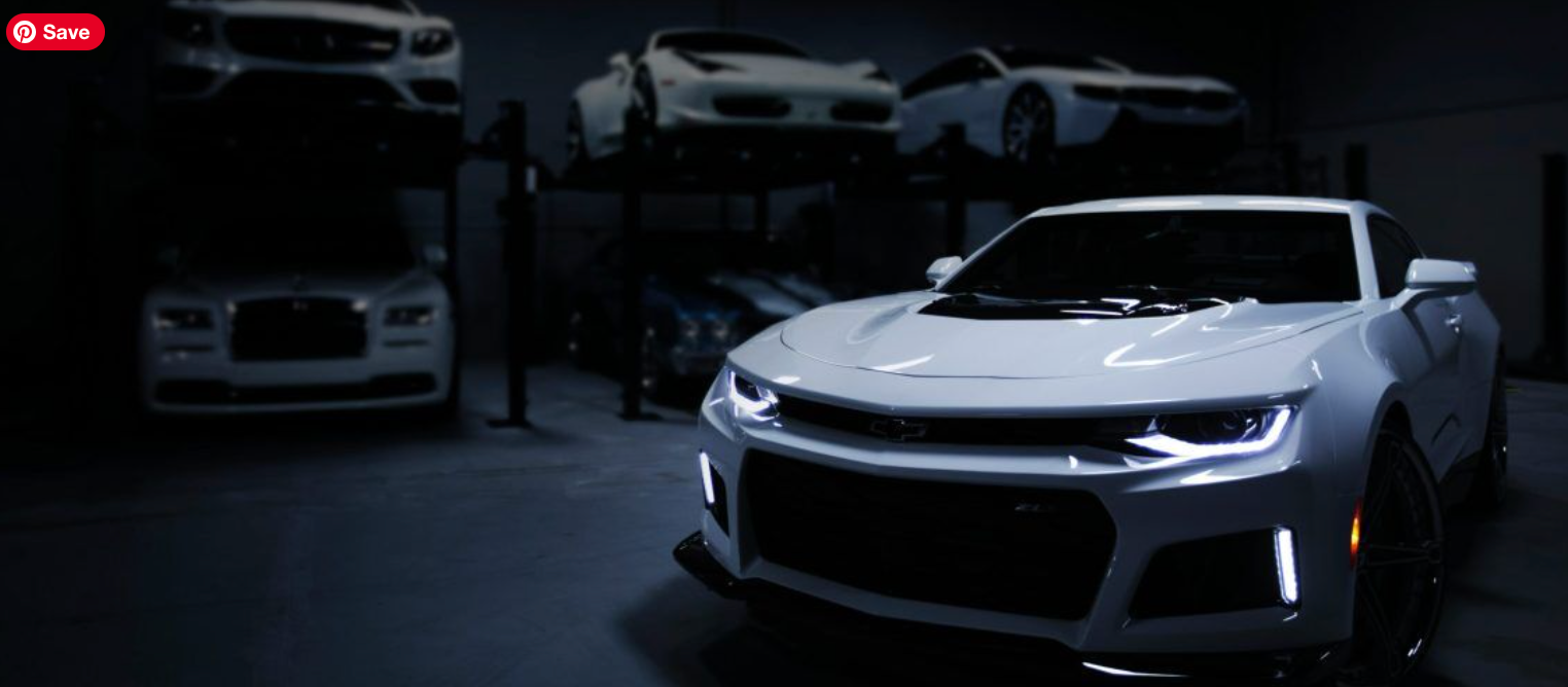 White Chevrolet Camaro parked in a garage with other luxury vehicles on a lift.