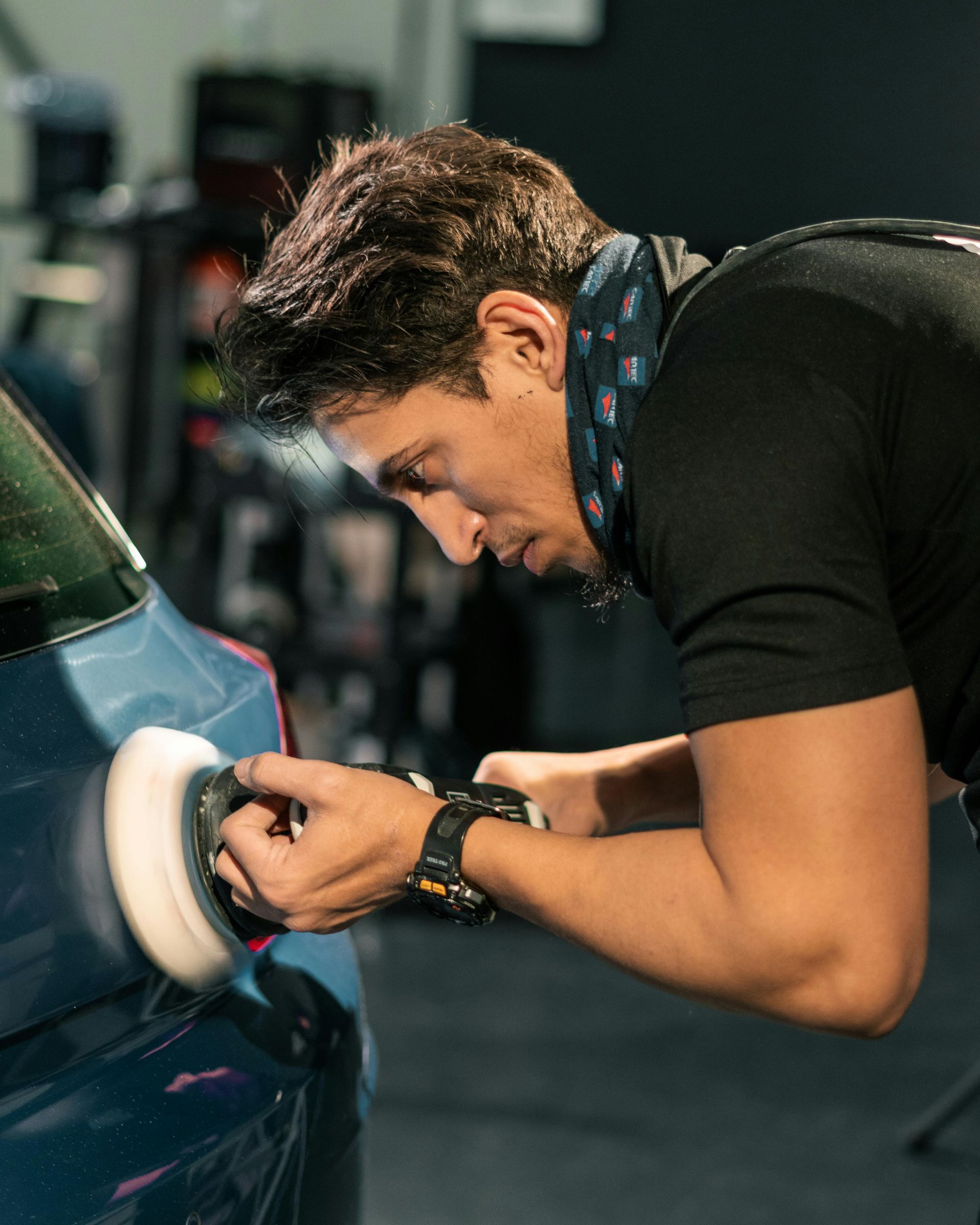Man polishing a blue car with a buffer in a shop setting.