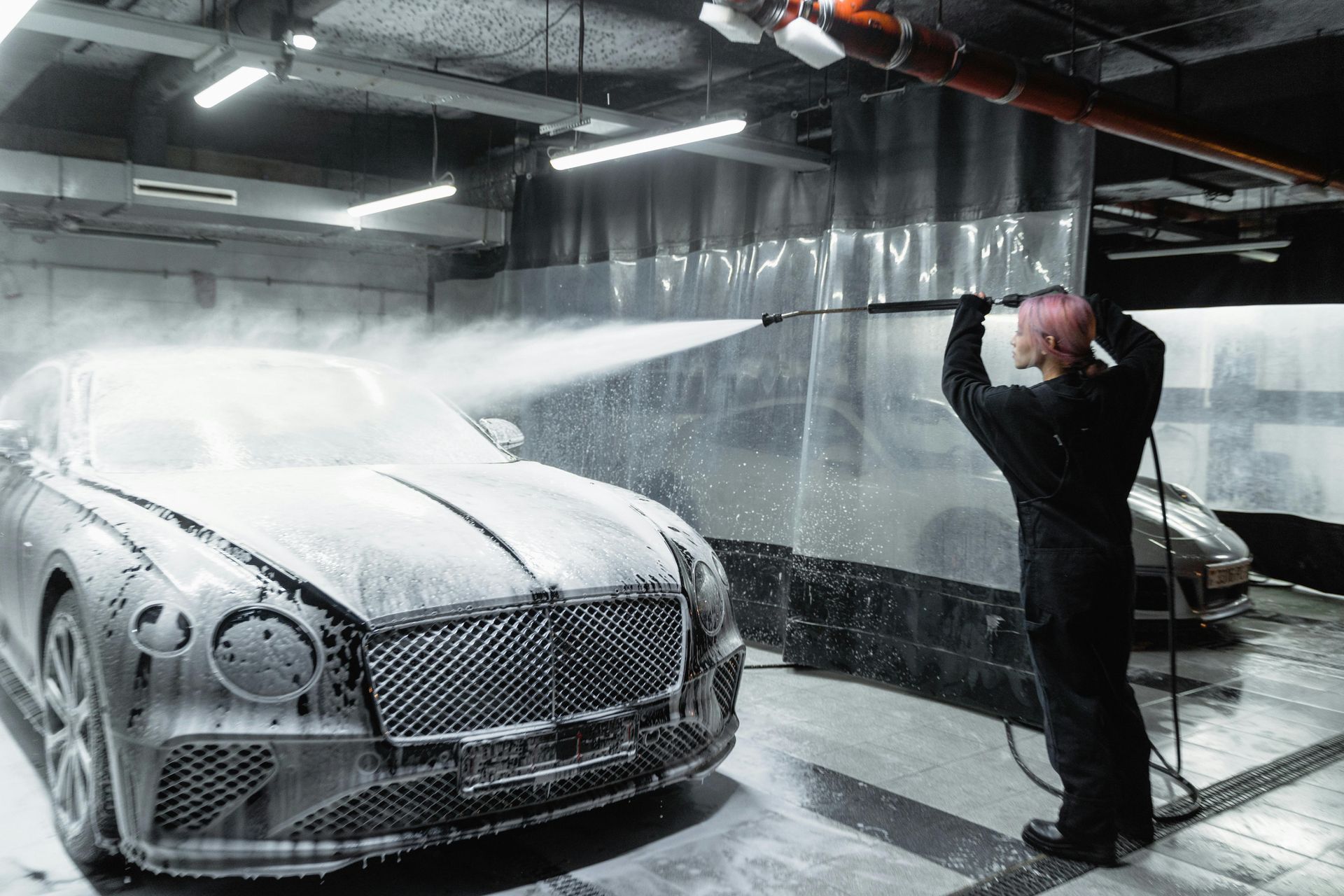 Person washing a silver Bentley with foamy soap in a car wash bay.