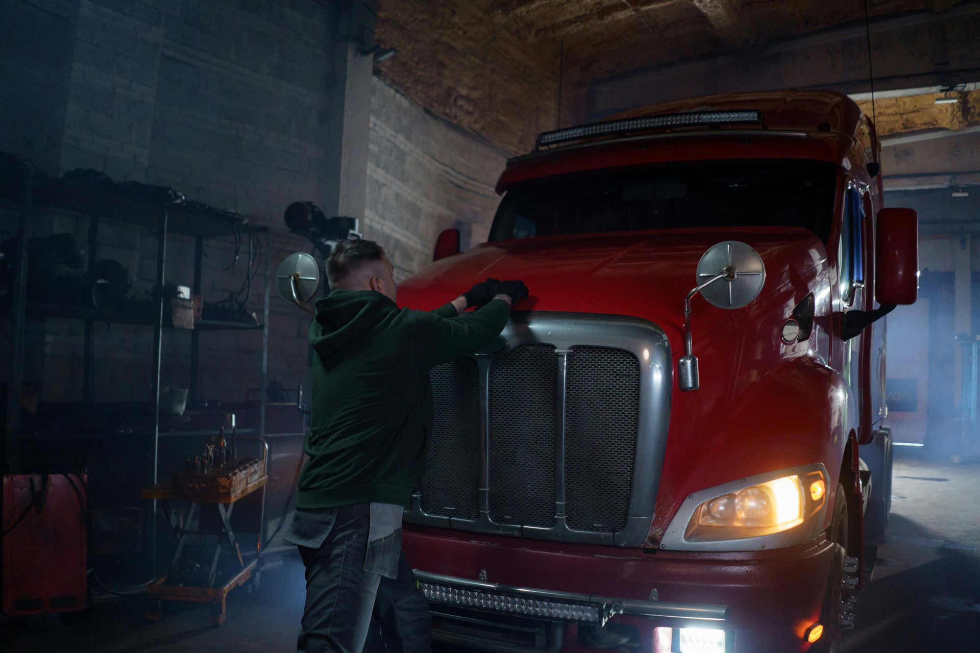 Man in green jacket working on the hood of a red semi-truck in a garage.