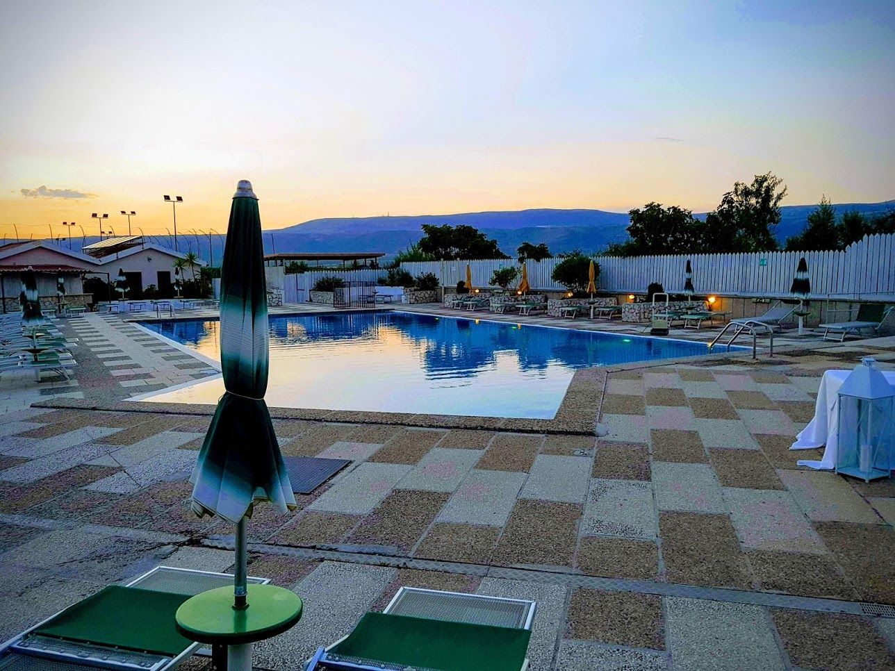 Terrazza a bordo piscina al tramonto con lettini, ombrellone e una tranquilla vasca riflettente che si affaccia sulle colline in lontananza.