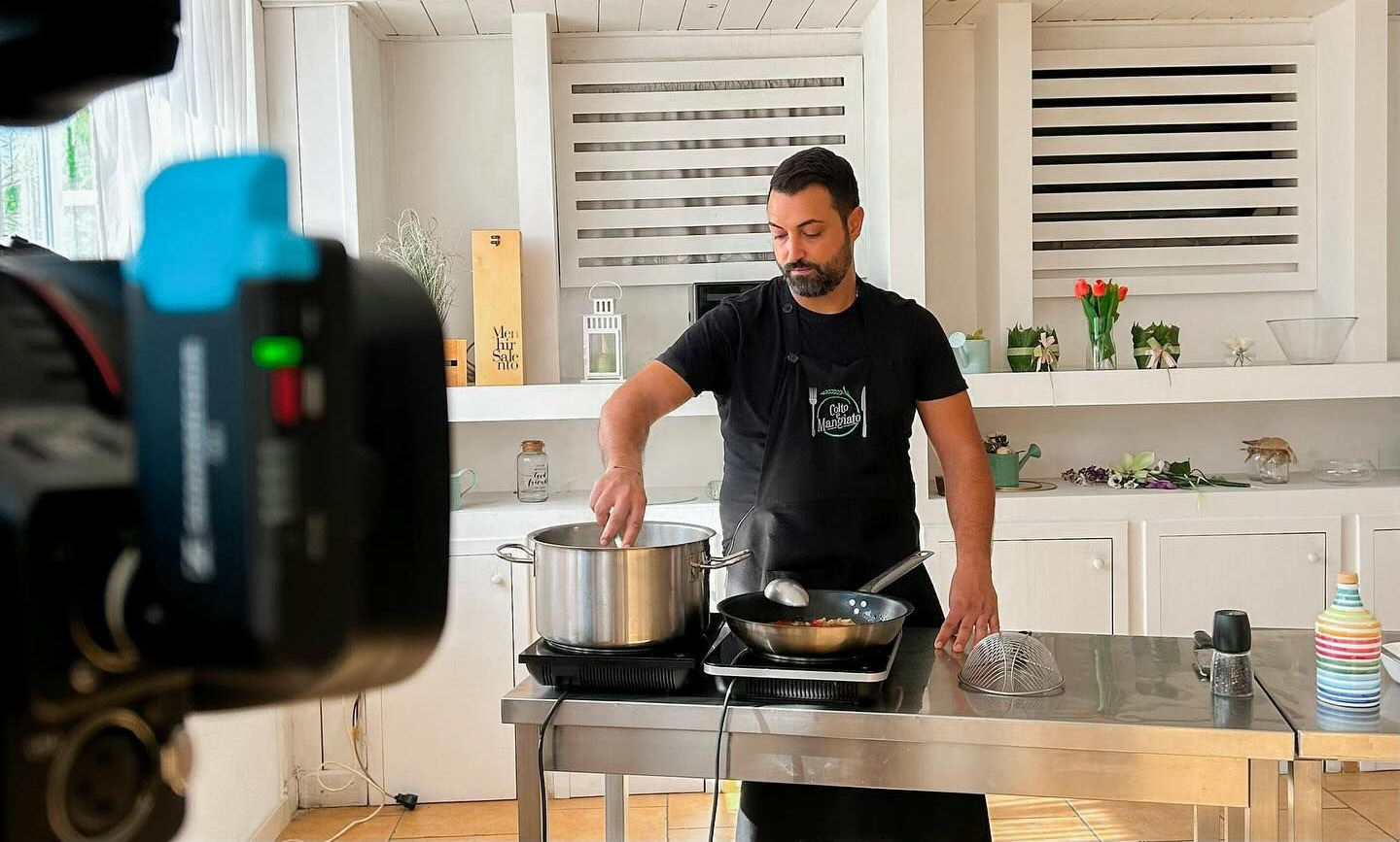 Un uomo cucina ai fornelli in una cucina luminosa, mescolando il contenuto di una pentola accanto a una padella e fiori sul bancone.