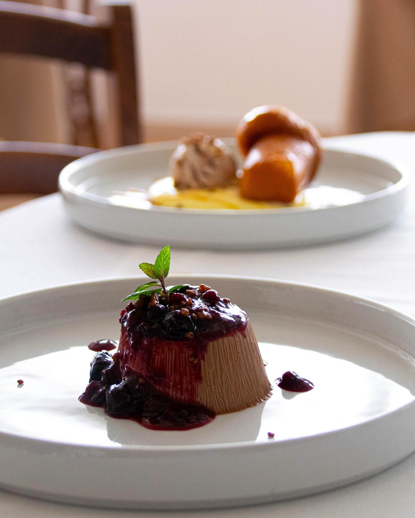 Due dessert impiattati su un tavolo, uno con salsa di frutti di bosco scuri e guarnizione di menta in primo piano.
