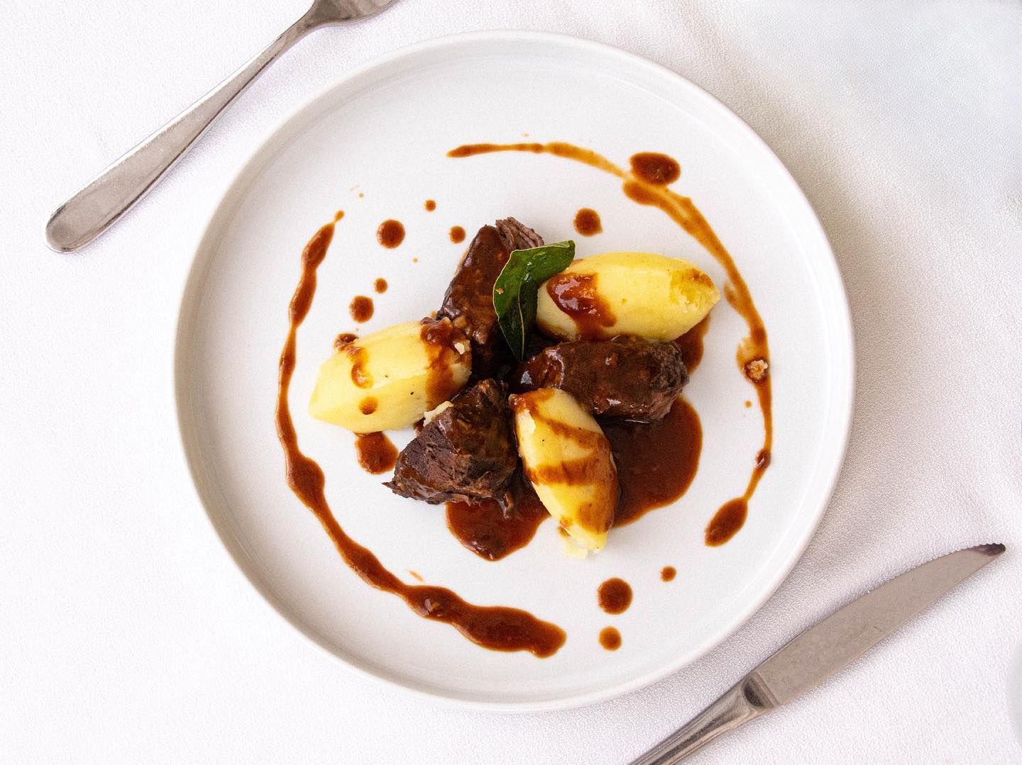 Dessert gourmet impiattato con vortici di salsa, guarnizione di frutta e cucchiaio su un tavolo bianco.
