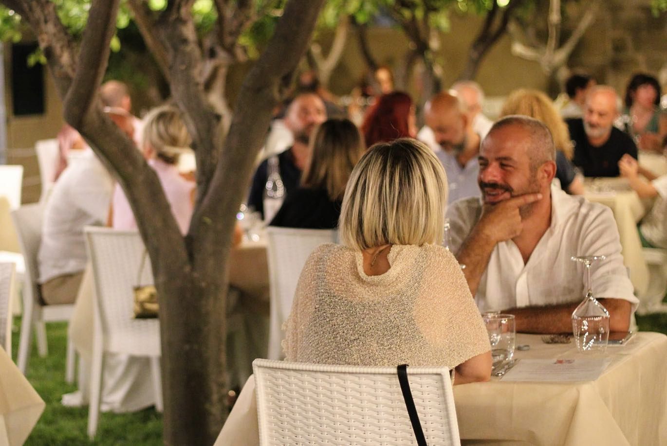 Una coppia cena in un ristorante all'aperto, seduta a un tavolo sotto gli alberi, con altri avventori sullo sfondo.