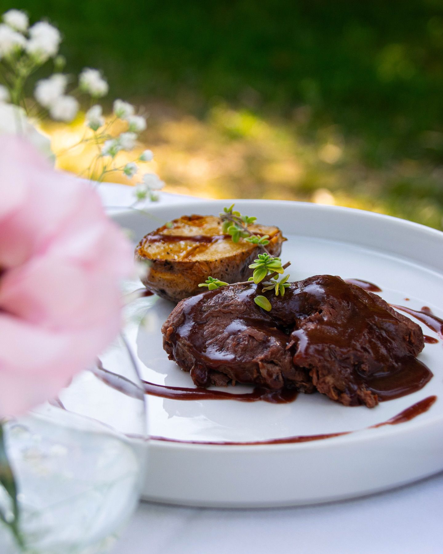 Bistecca alla griglia su un piatto bianco con patate arrosto e salsa, all'aperto su un tavolo con fiori nelle vicinanze.