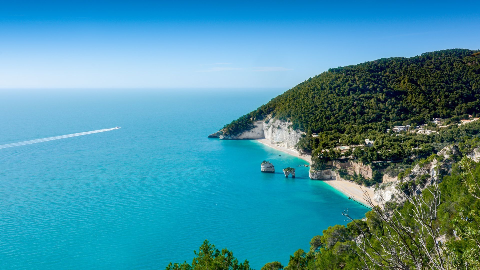 Mare turchese accanto a una costa rocciosa e verdeggiante con una piccola spiaggia e barche sotto un cielo azzurro limpido.