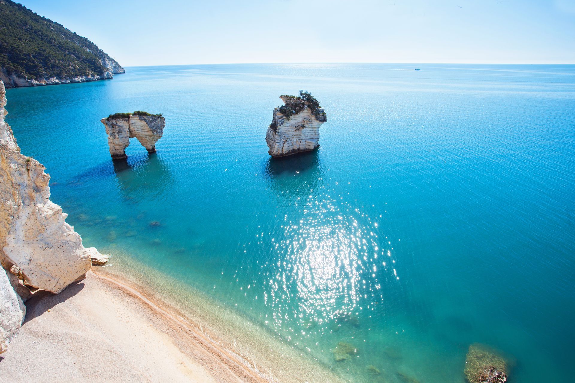 Mare turchese illuminato dal sole con due archi di roccia naturali accanto a una spiaggia sabbiosa e costa a picco sul mare.