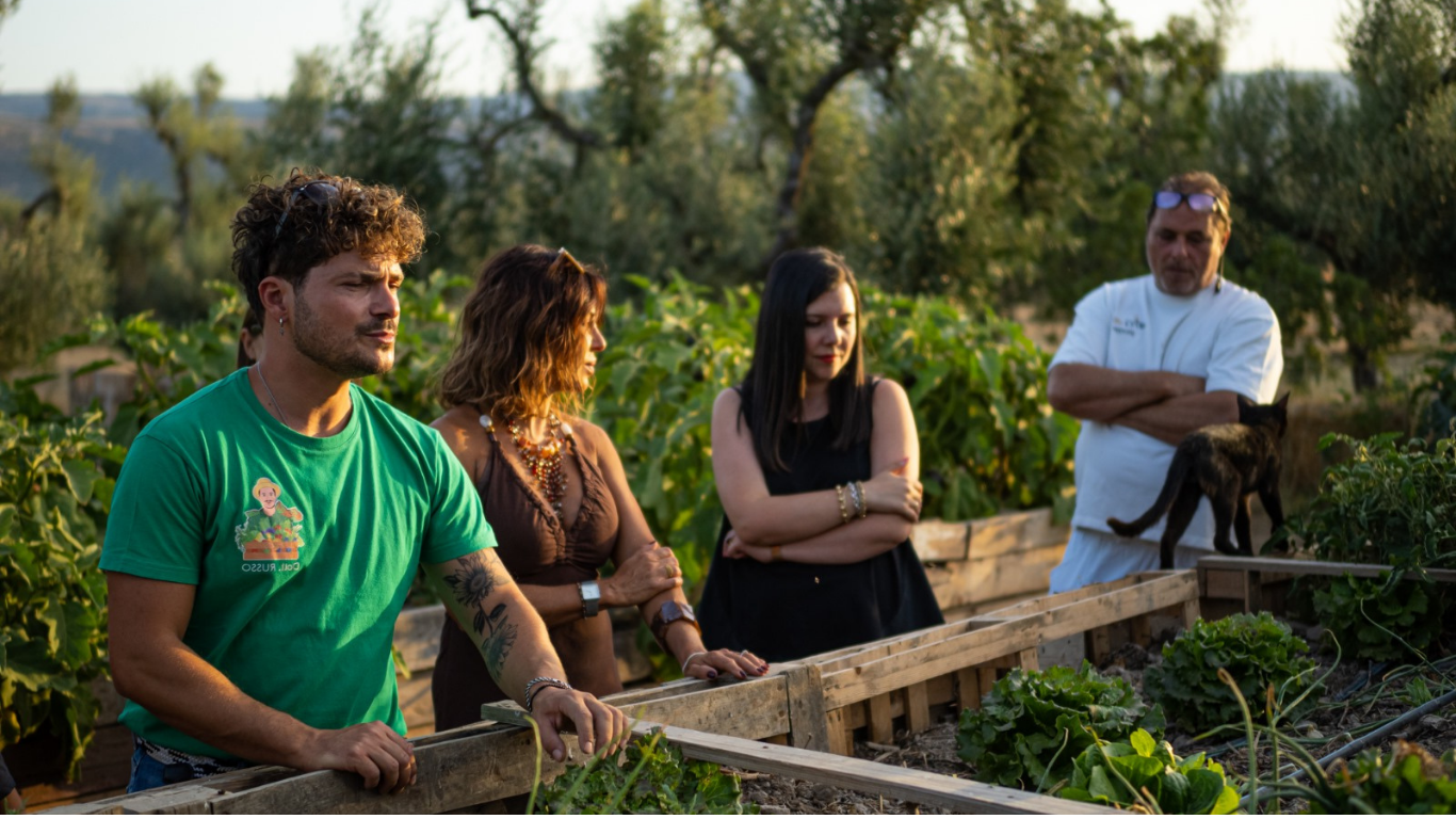 Persone in piedi che lavorano in un giardino accanto a aiuole rialzate in un campo soleggiato all'aperto.