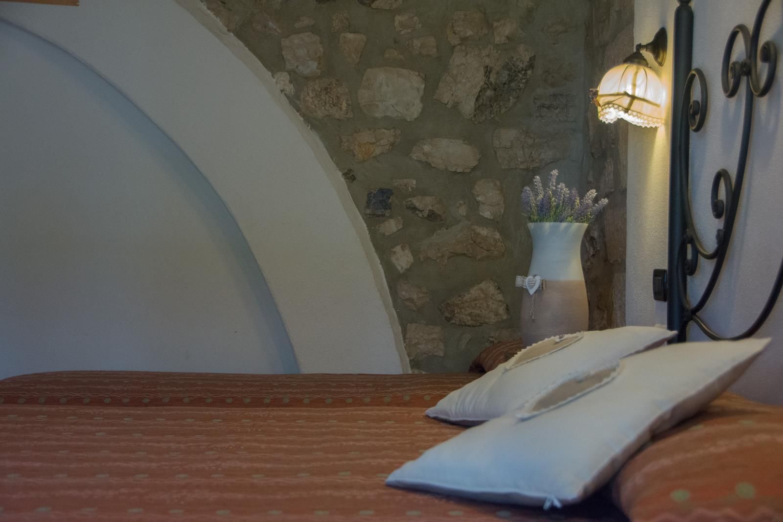 Accogliente angolo camera da letto con parete in pietra, nicchia ad arco, lampada e cuscini su un letto.