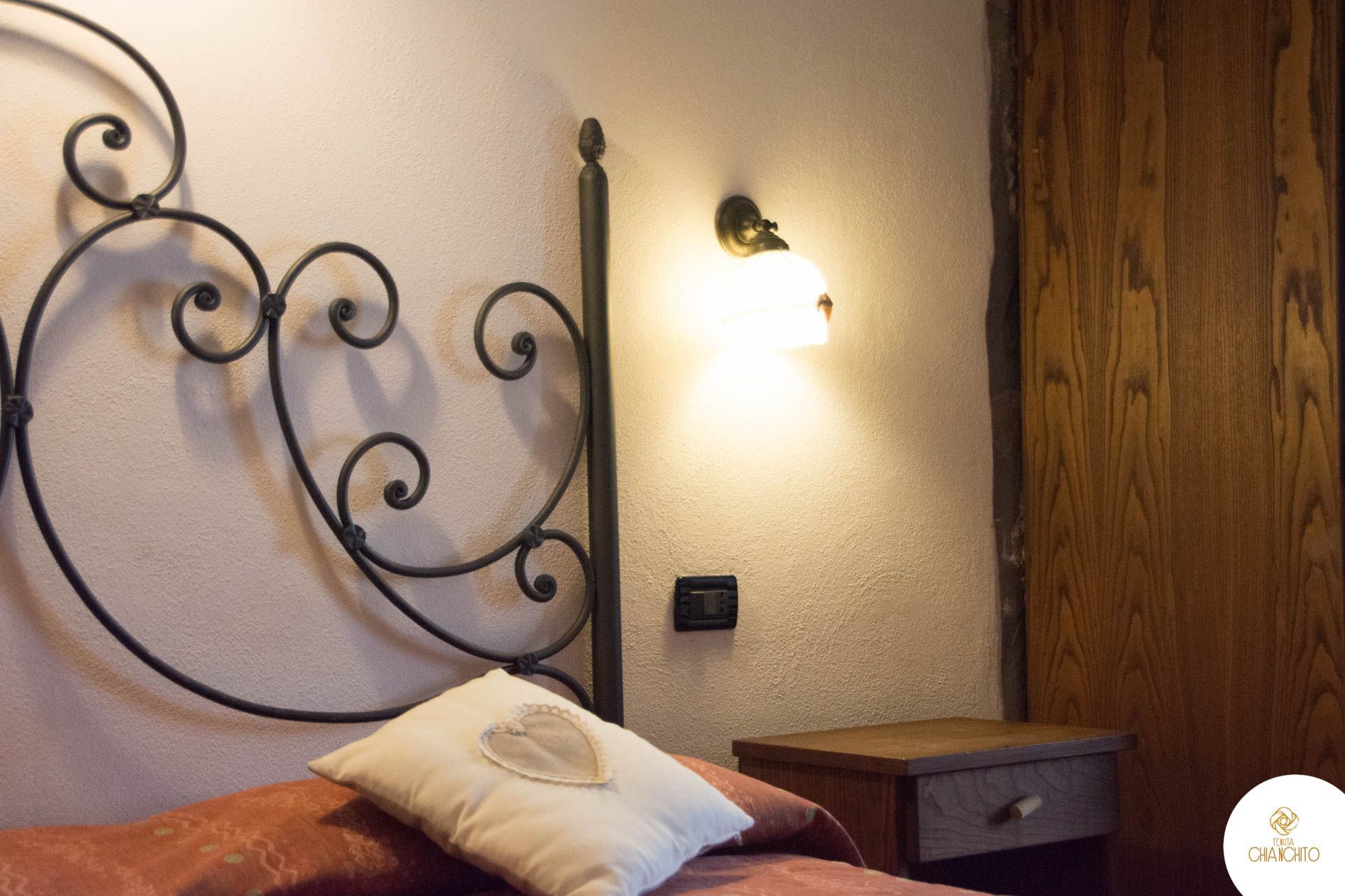 Camera da letto con testiera in ferro battuto, lampada da comodino, comodino in legno e cuscini decorati.