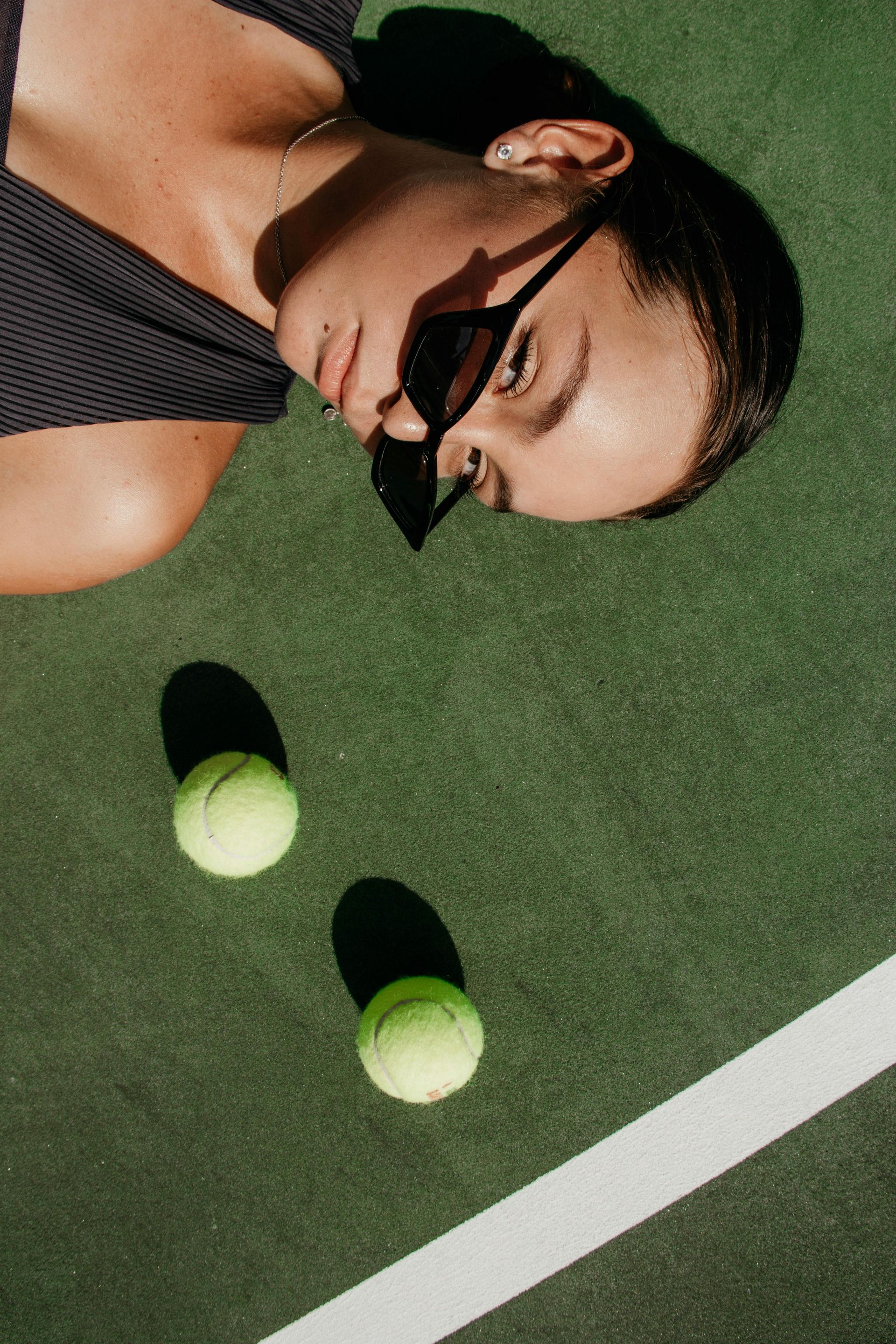 Una persona è sdraiata su un campo da tennis verde accanto a due palline da tennis, con indosso una mascherina nera.