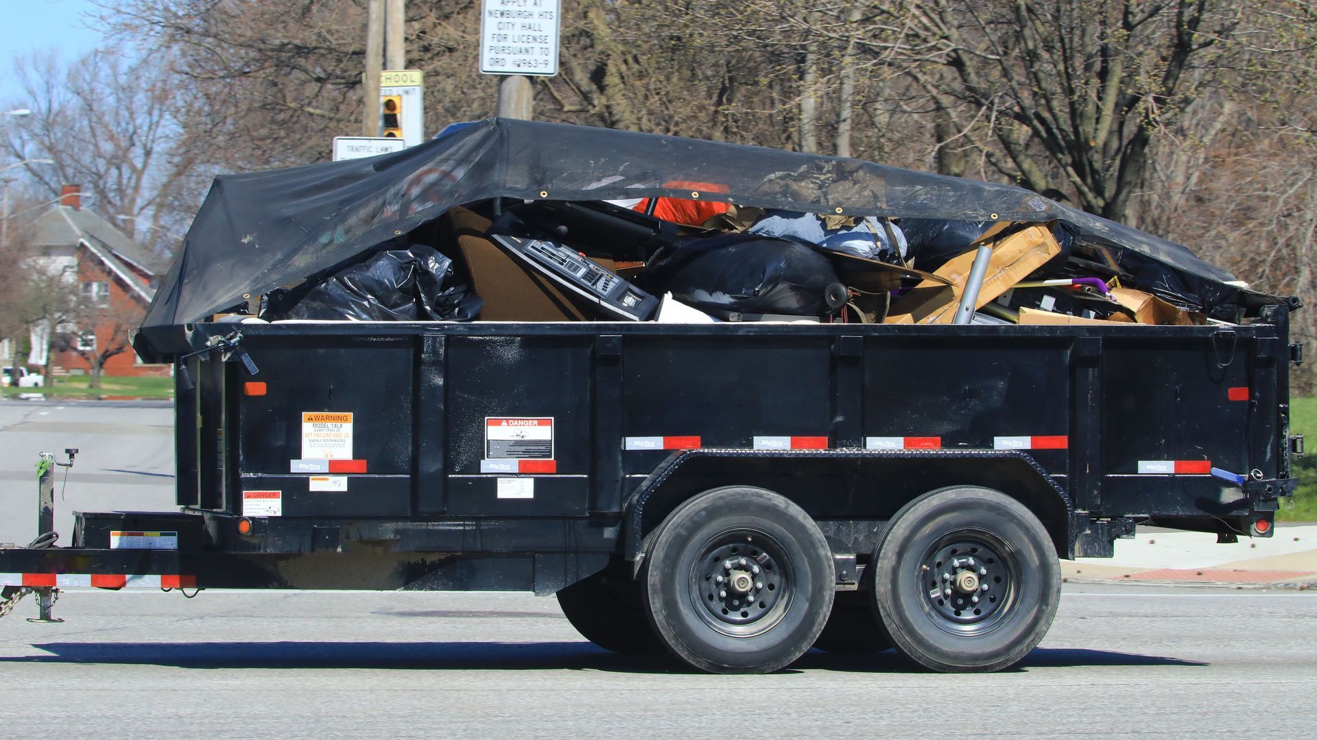 Black trailer filled with trash and covered by a black tarp, parked on a street.