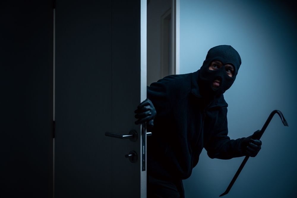 A person in a black balaclava and gloves holding a crowbar peeks through a slightly open door in a dimly lit room.