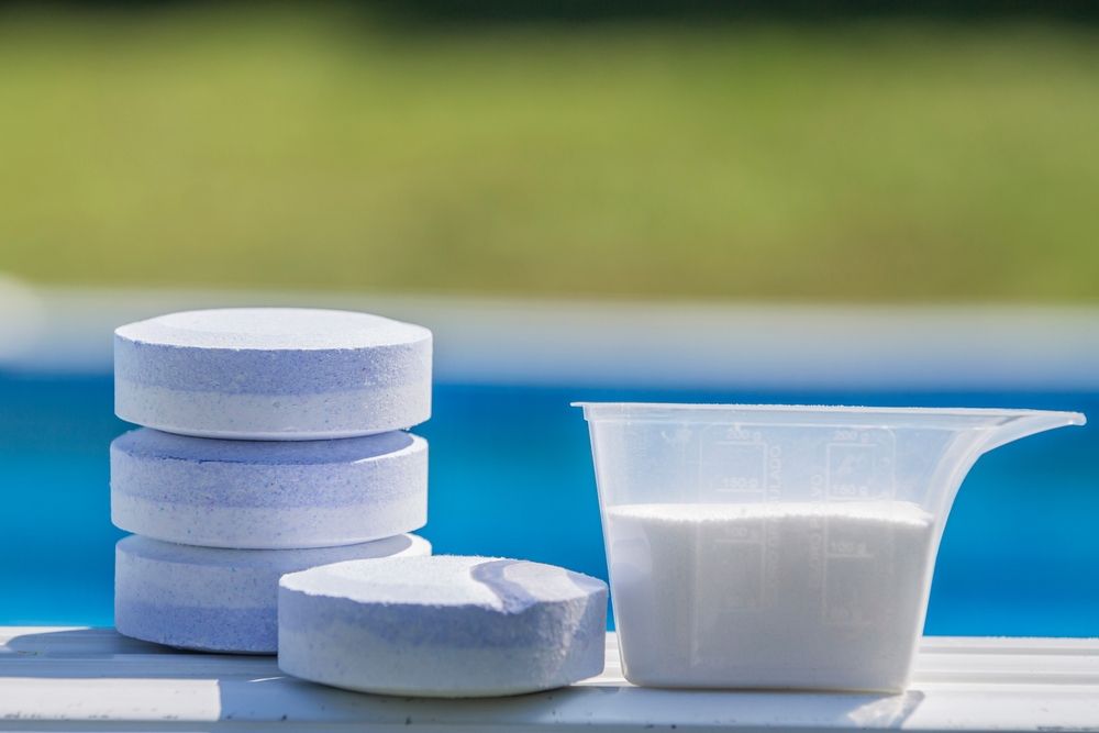 A stack and a single pile of white and blue pool chlorine tablets sit next to a measuring cup of white chemical powder.