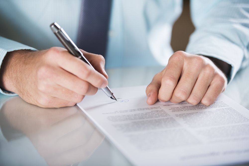A person in a light blue shirt signing a document with a silver pen at a desk.