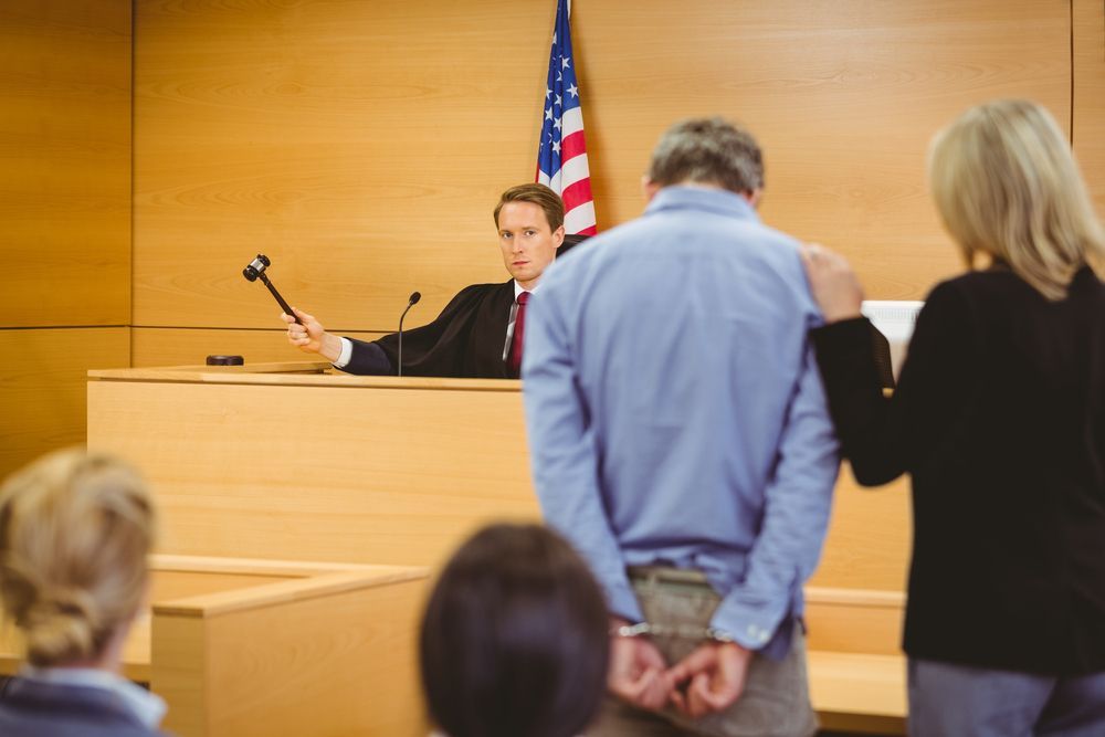 A judge in a courtroom holds a gavel while a person in handcuffs stands with a woman in front of the judge's bench.