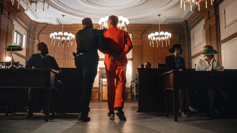 A law enforcement officer leads a person in an orange jumpsuit through a courtroom, walking toward the back of the room.