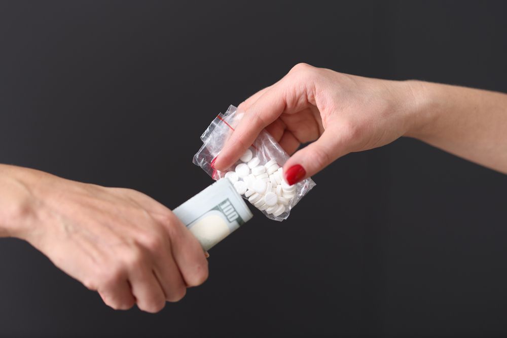 One hand exchanges a roll of hundred-dollar bills for a plastic bag filled with white pills against a black background.