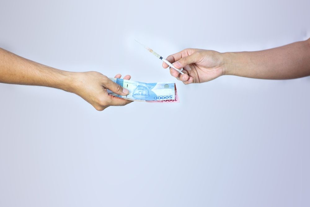 Two hands exchange a syringe for a stack of Indonesian Rupiah banknotes against a plain white background.
