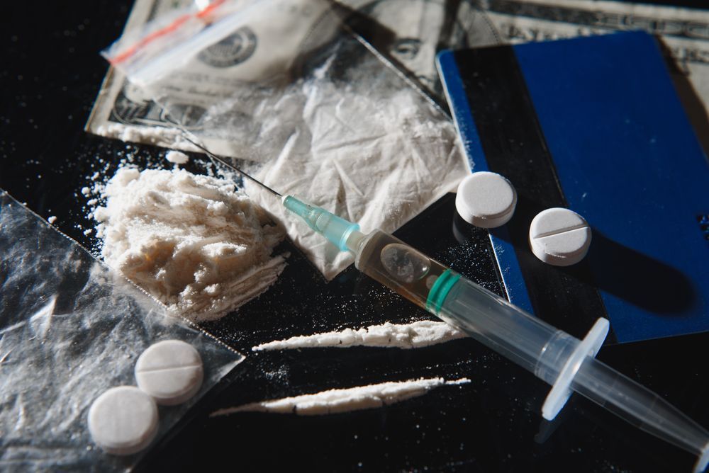 A syringe, white powder, pills, a credit card, and US currency on a dark surface.