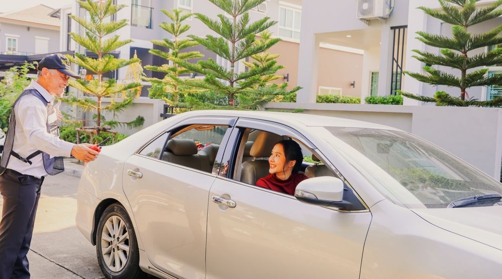 A security guard in a uniform speaks to a driver inside a parked silver sedan in a residential neighborhood.