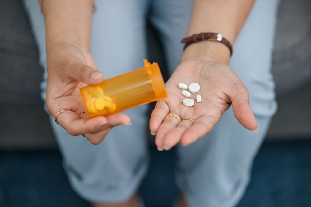 A person holds an orange prescription bottle and several white pills in their open hand.