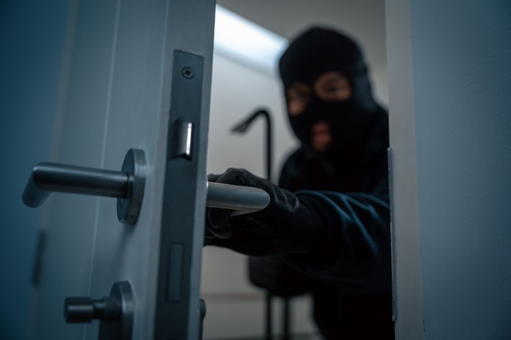 A person in a black balaclava and gloves holding a crowbar as they open a door.