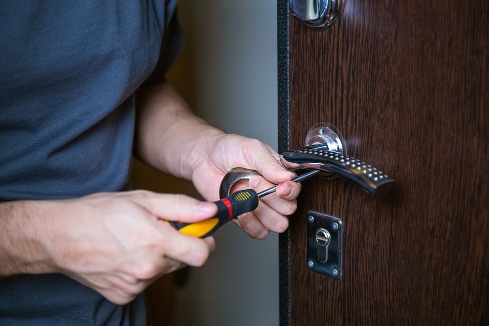 A person in a gray shirt uses a screwdriver to tighten or remove a screw on a dark wood door's handle mechanism.