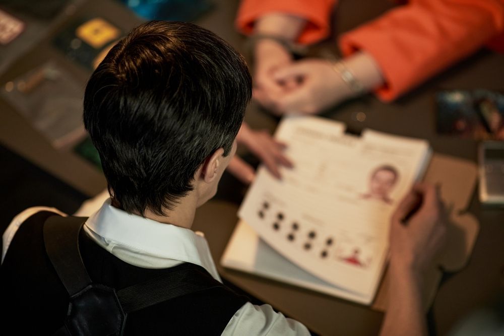 An individual in a white shirt and vest holds a file with images, while hands in orange sleeves rest on the table nearby.