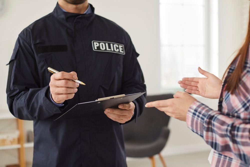A police officer in a dark uniform writes on a clipboard while a person in a plaid shirt gestures during a conversation.