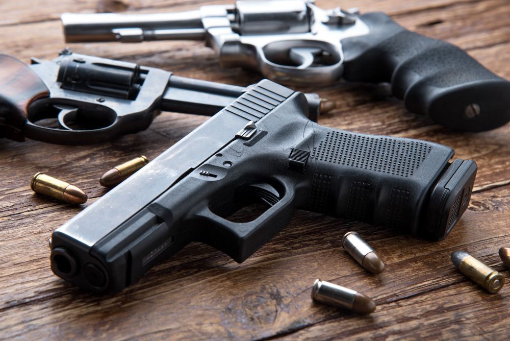 Three handguns, including a black semi-automatic pistol and two revolvers, arranged on a rustic wooden table with bullets.