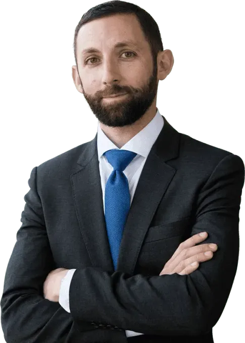 A person with a beard, wearing a dark suit and a blue tie, stands with arms crossed against a white background.