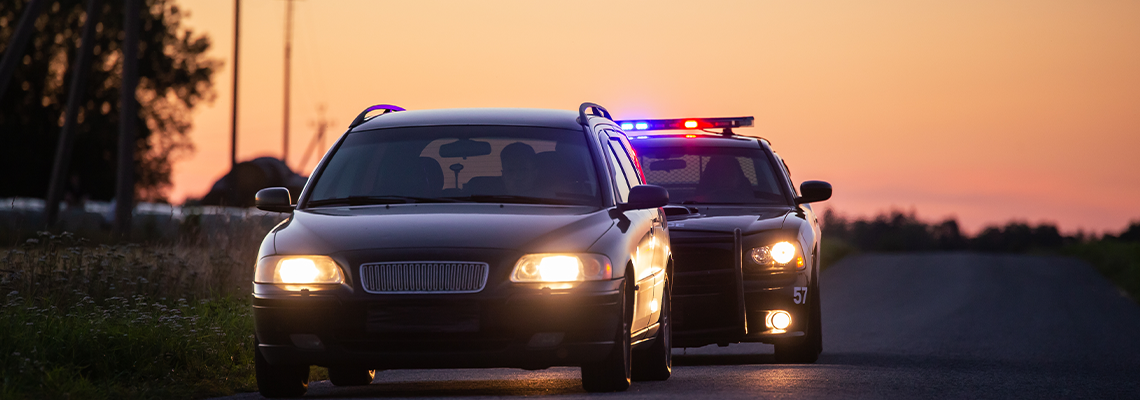 A police cruiser with flashing lights pulls over a dark car on a rural road at sunset.