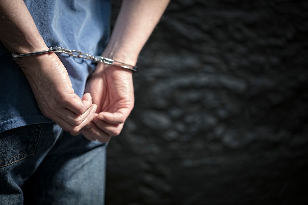 A person wearing a blue shirt and jeans with their hands behind their back in handcuffs against a dark, textured wall.