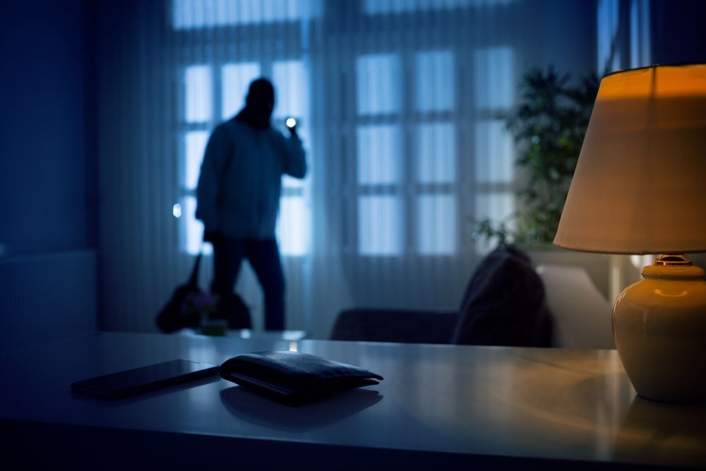 A shadowed figure with a flashlight enters a dimly lit room, with a wallet and phone resting on a table in the foreground.