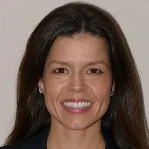 Woman with brown hair, smiling, wearing earrings and a dark blazer.