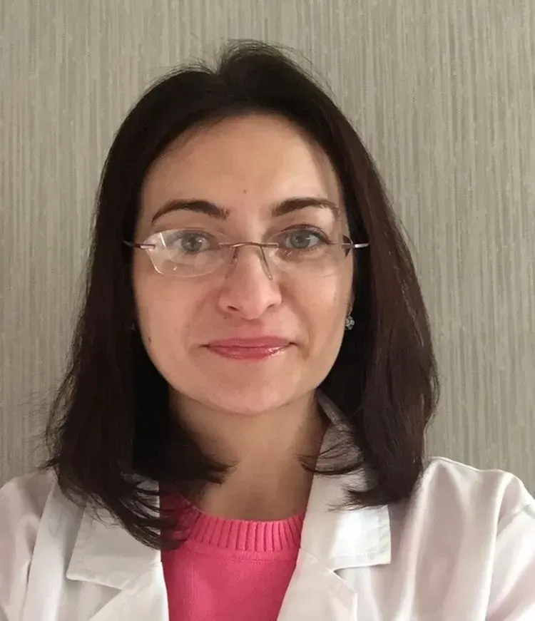 Woman in glasses and white coat, looking directly at the camera. Pink sweater visible, neutral background.