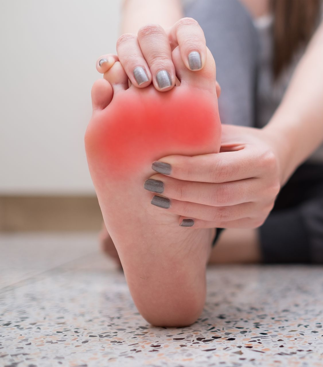 Woman holding her foot, a red area indicating pain on the sole, indoors.
