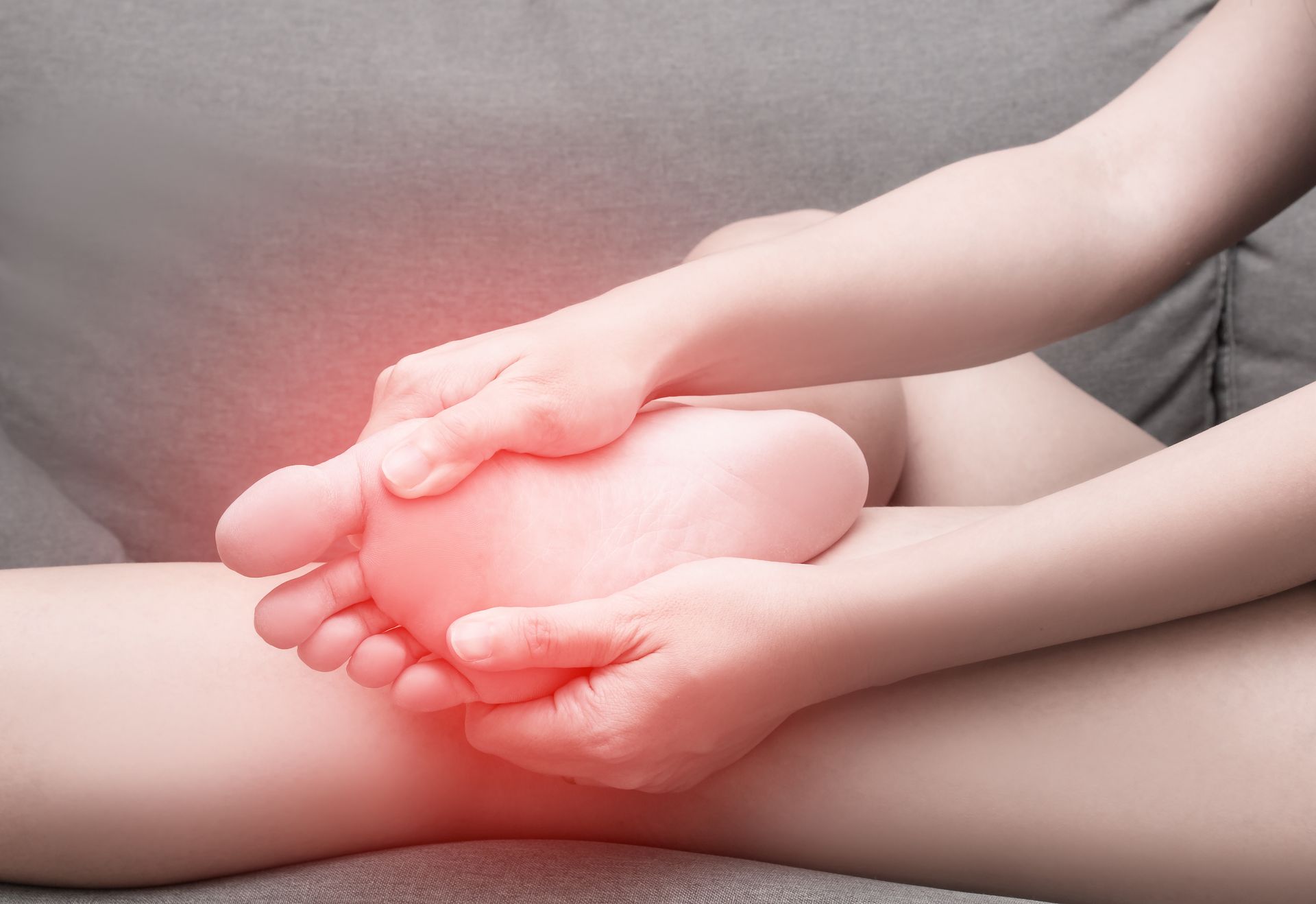Person's hands holding their foot, highlighting the sole. Red glow indicates pain. Sitting on a gray surface.