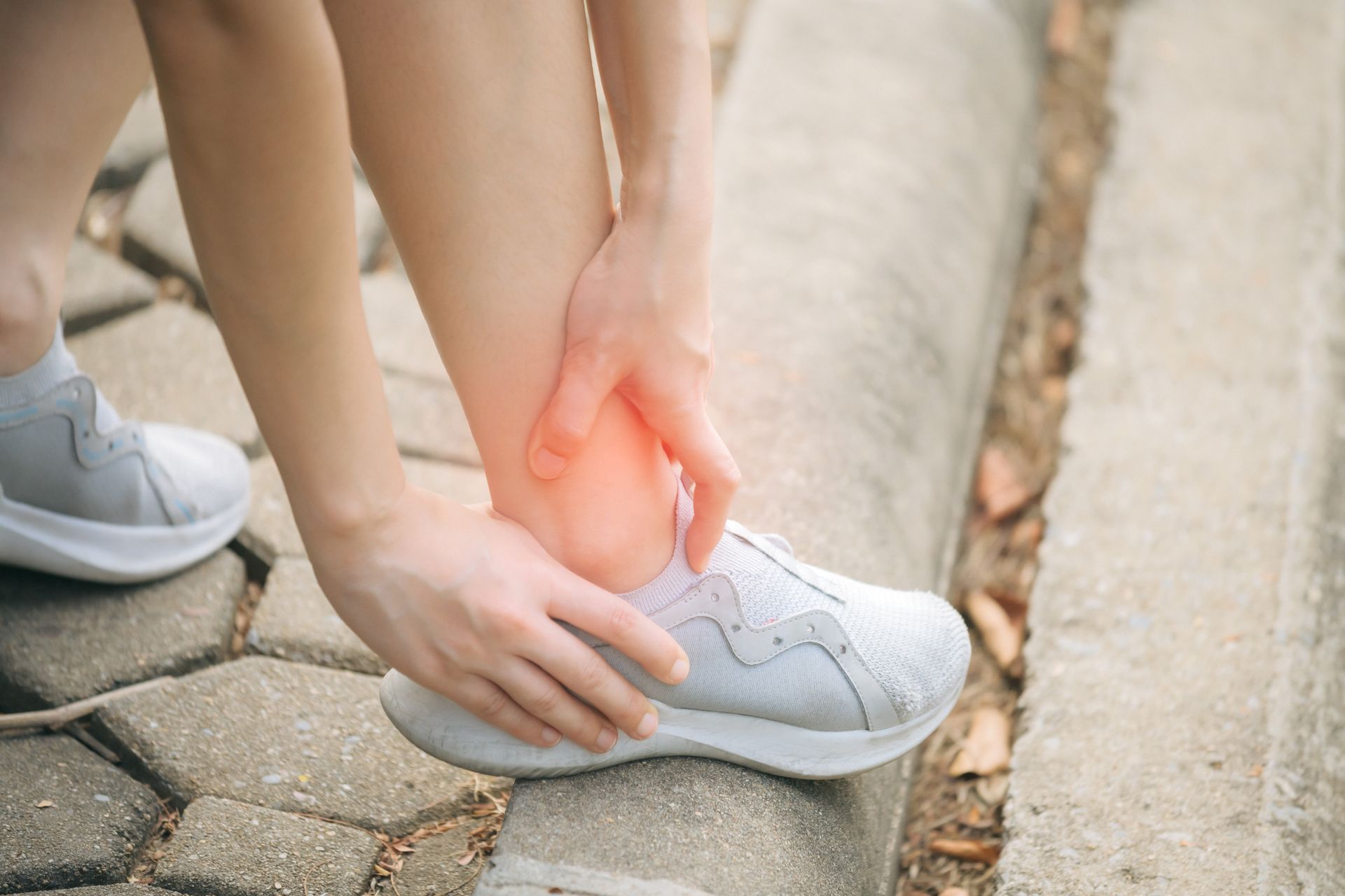 Person holding their ankle in pain while wearing a grey shoe, red highlight indicating pain. Outdoors.