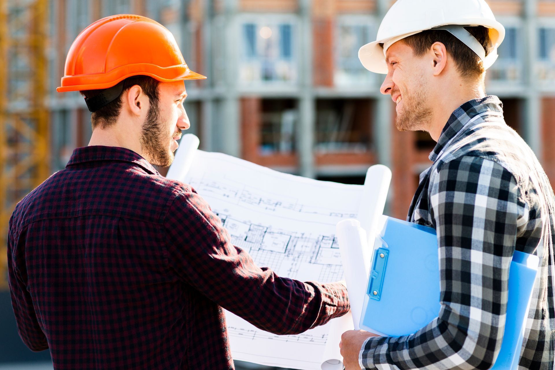 Deux ouvriers du bâtiment, casqués, examinent des plans sur un chantier.