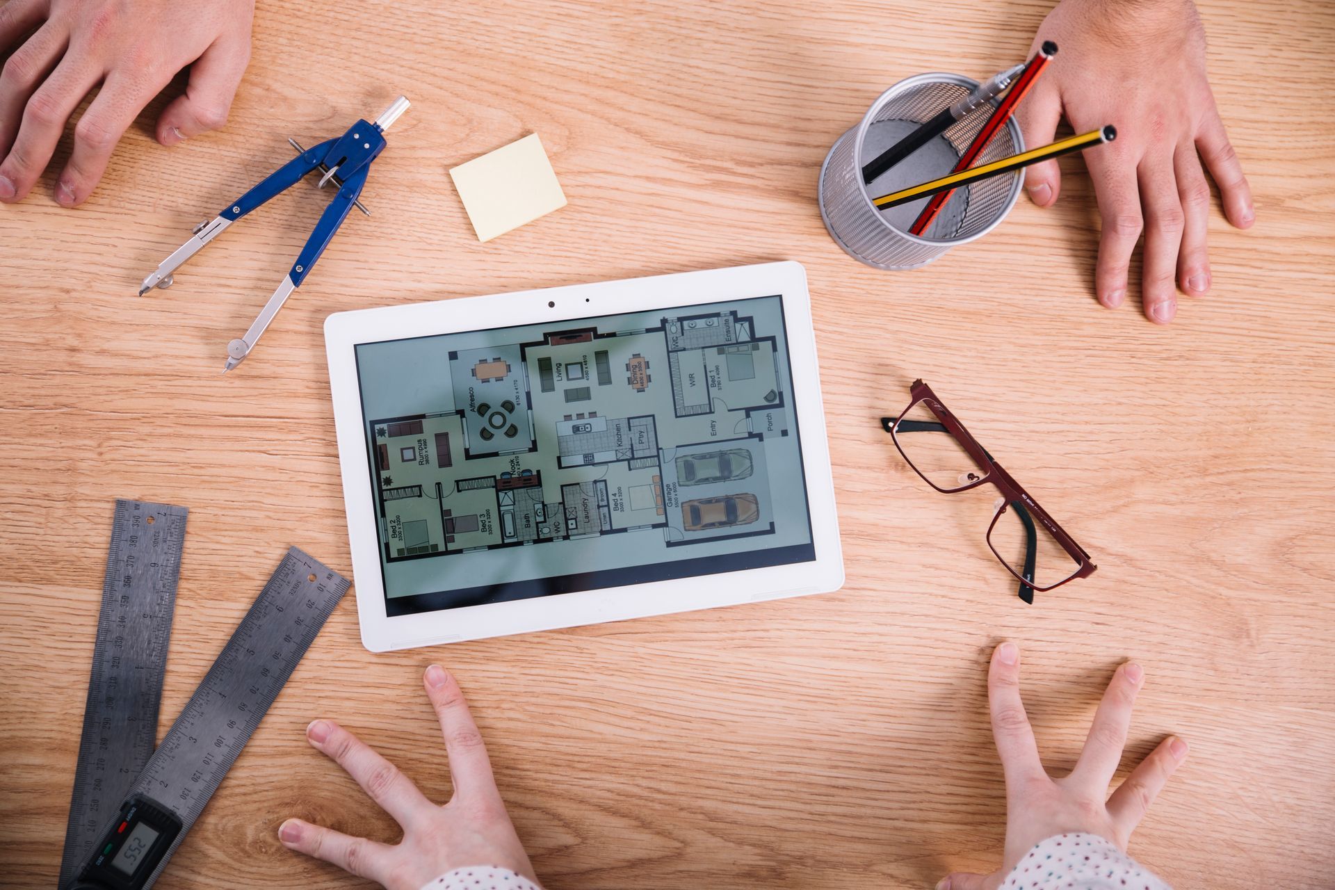 Des mains autour d'une tablette affichant un plan, entourées d'outils : compas, crayons, règle, lunettes et post-it sur une table en bois.