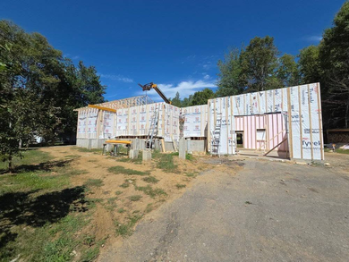 Construction en cours : murs extérieurs à ossature bois, recouverts de revêtement, posés sur des fondations en béton, ciel bleu, arbres en arrière-plan.