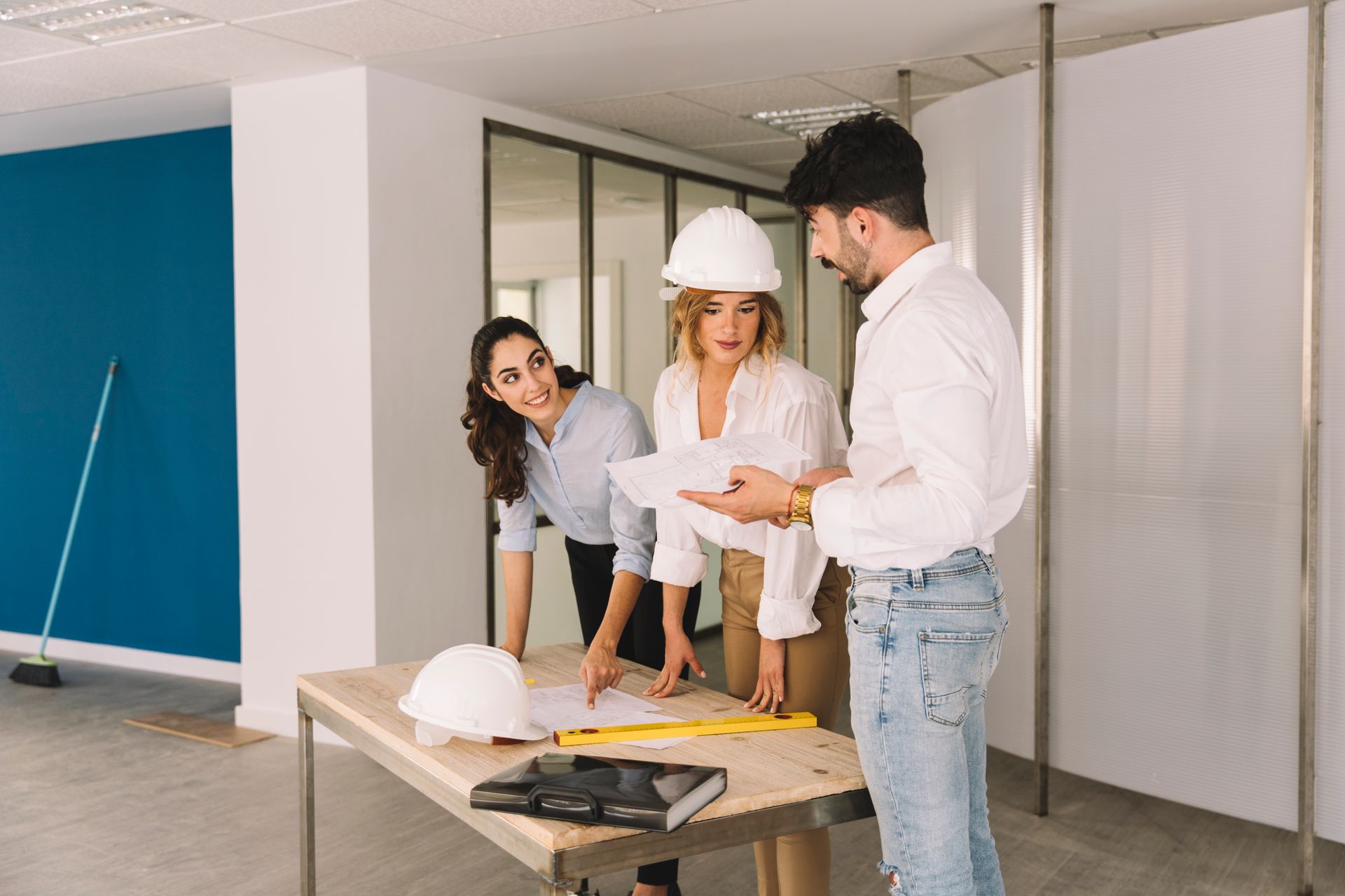 Trois personnes dans une pièce, en train d'examiner des plans. L'une d'elles porte un casque de chantier ; toutes sourient et discutent des projets autour d'une table.