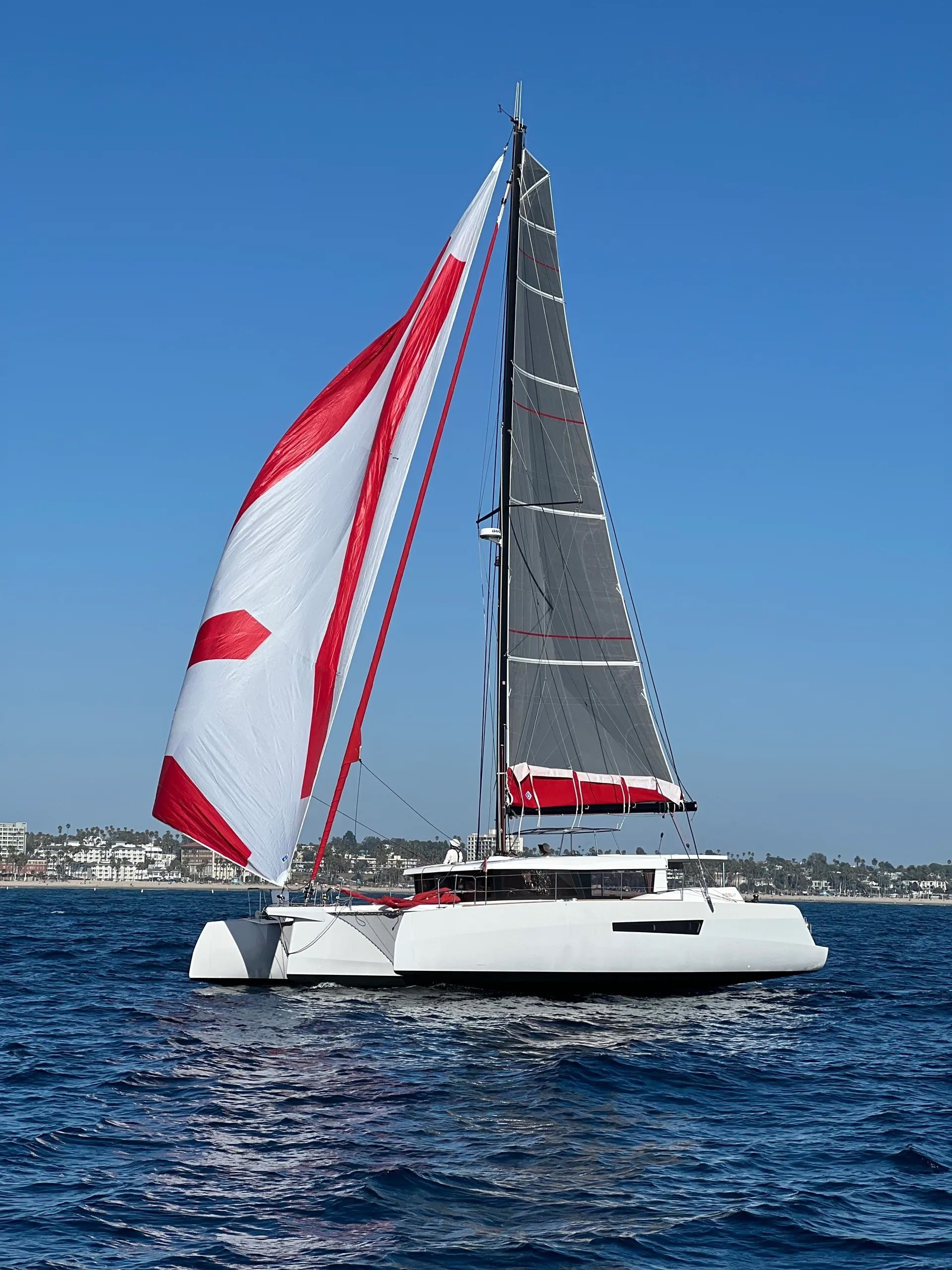 White catamaran sailboat with red-and-white sails on blue water under a clear sky