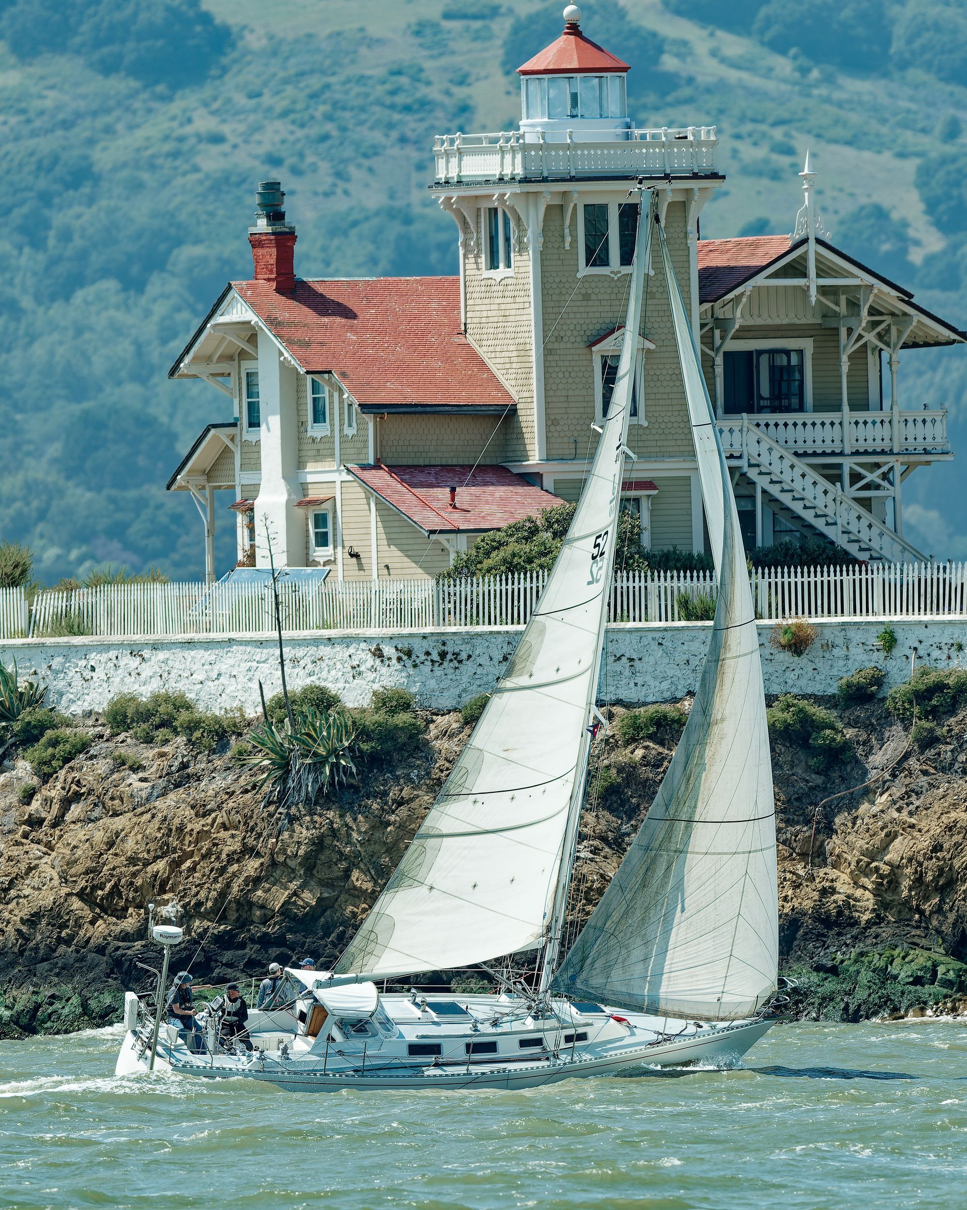 Sailboat in choppy water beside a white lighthouse and house with red roofs on a rocky shoreline