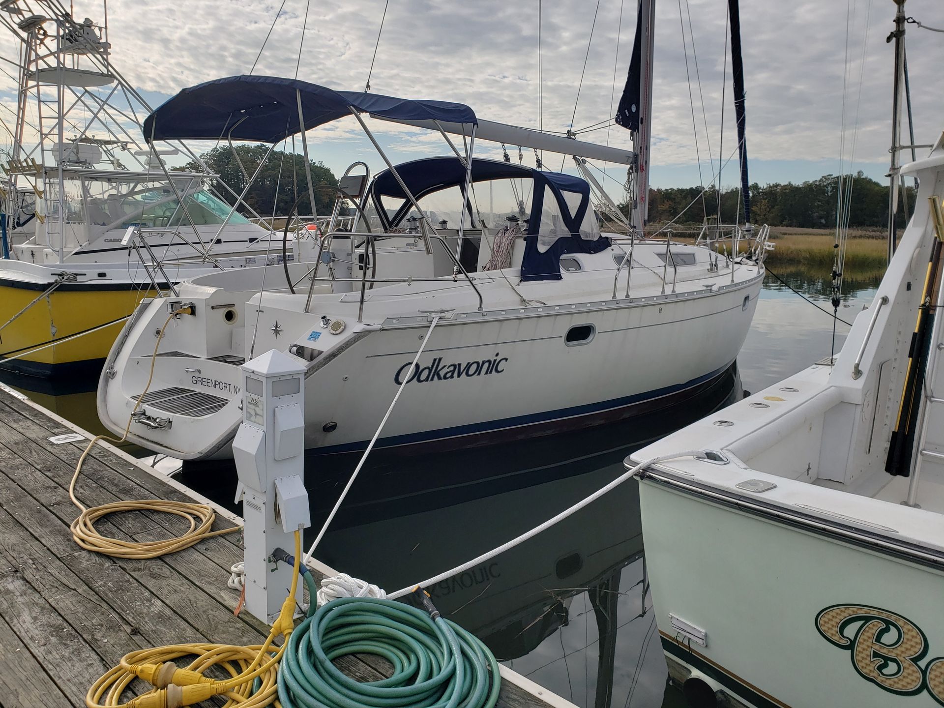 Sailboats docked at a marina, with a white sailboat in the center and a green hose on the pier.