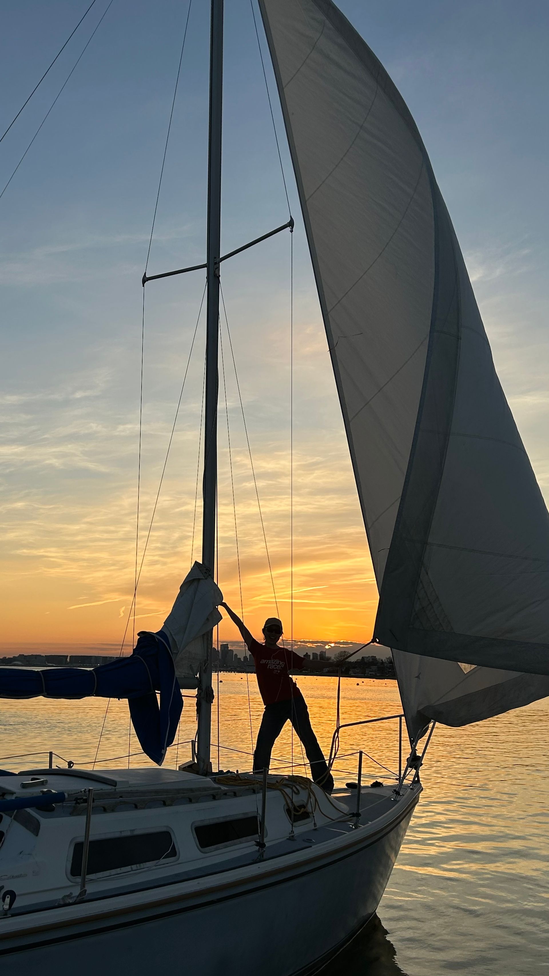 Sailboat at sunset with two people adjusting the sails on calm water.