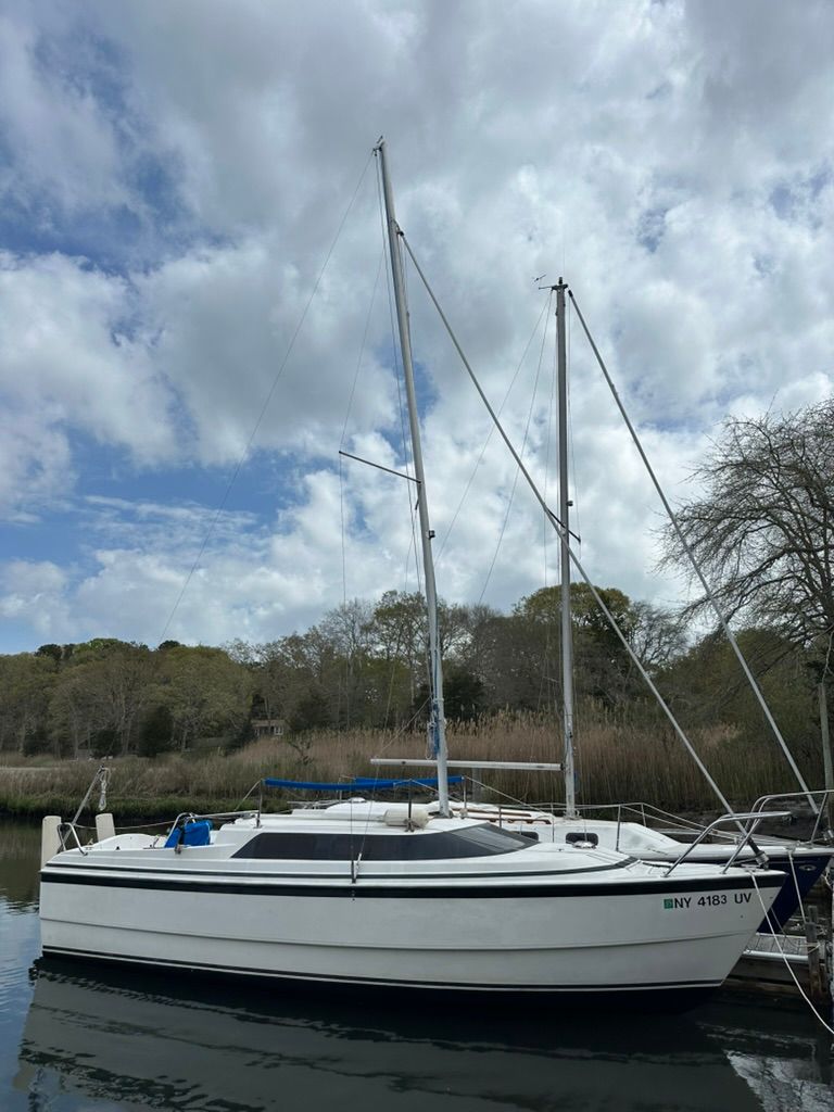 White sailboat moored on calm water under cloudy sky, with trees and shoreline in the background